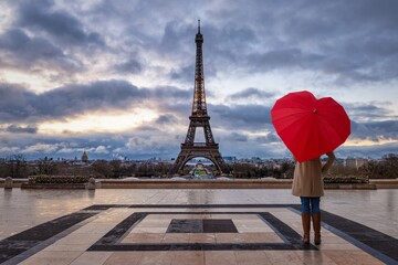 A woman with a red, heart shaped umbrella looks at the famous Eiffel Tower of Paris, France, during a rainy, moody day