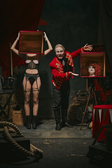 Tricks. Magician in red coat showing tricks with headless female assistant holding boxes over dark...