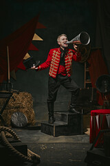 Ringmaster, magician in red coat shouting into megaphone over dark retro circus backstage background. Announcement of show. Concept of circus, theater, performance, show, retro and vintage © master1305