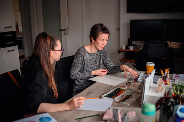 Two women share a creative moment; one chooses paper while the other prepares to paint, a symbol of...