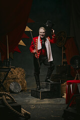 Man with pale face makeup, in red stage costume and hat making creative performance over dark retro circus backstage background. Concept of circus, theater, performance, show, retro and vintage © master1305