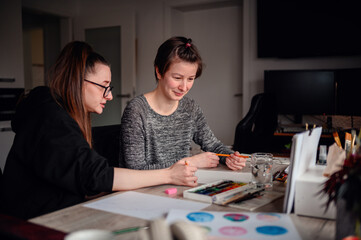 Fototapeta premium In a cozy, cluttered workspace, two women share a light-hearted moment over their art projects, surrounded by creative tools