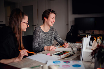 In a cozy, cluttered workspace, two women share a light-hearted moment over their art projects,...