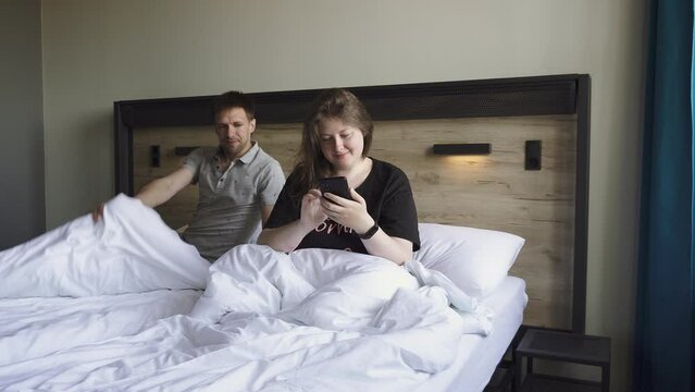Woman is watching videos and photos on smartphone in bed when man comes to her at home. Happy white married couple looking at phone with smile before going to sleep in bedroom indoor.