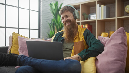A smiling bearded redhead man lounging indoors with a laptop and headphones enjoying leisure time.