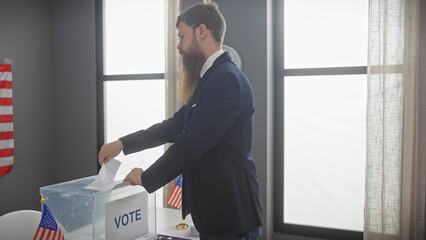 A man with a beard casting a vote in a ballot box inside a room with american flags.