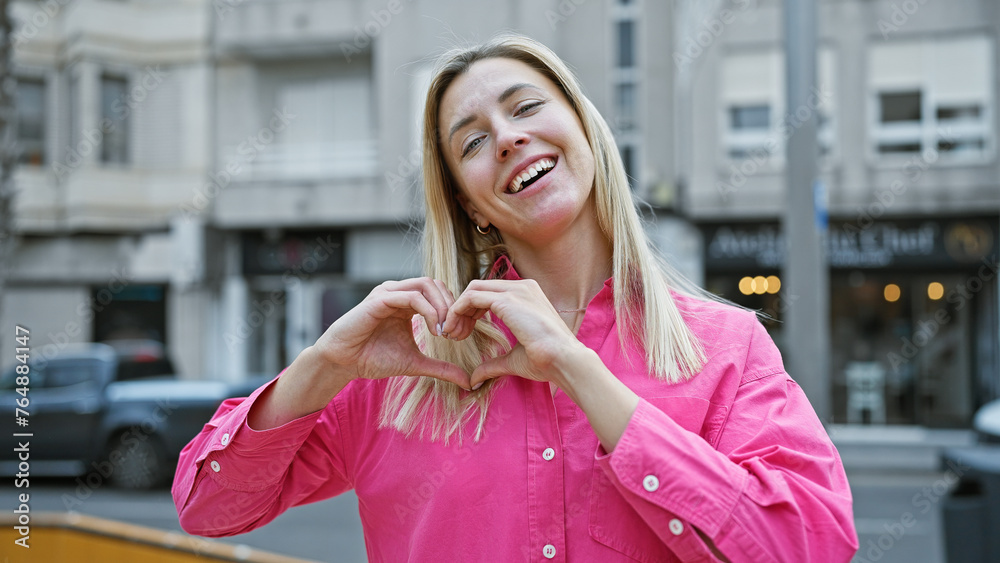 Poster smiling blonde woman forming a heart with her hands on a city street, conveying love and urban lifes