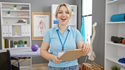 A cheerful young woman in a clinic room with medical posters, holding a clipboard and wearing a...
