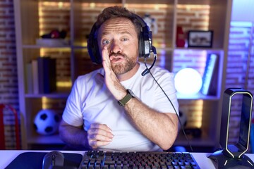 Middle age man with beard playing video games wearing headphones hand on mouth telling secret...