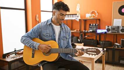 Handsome bearded hispanic man playing acoustic guitar in a cozy recording studio with microphone...