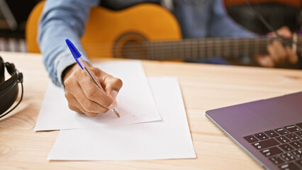 Hispanic man writing music studio notes with guitar and laptop