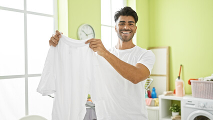 Handsome young man smiling while holding a white t-shirt in a bright laundry room.