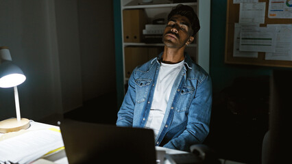 Tired hispanic man in office at night with computer and lamp, contemplating workload