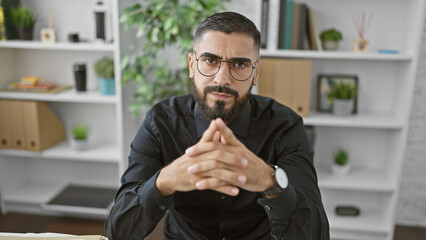 A confident young man with a beard sits in a modern office setting, exuding professionalism and...