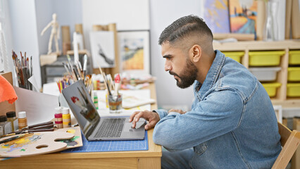 Handsome man with a beard working on a laptop in a creative studio surrounded by art supplies.