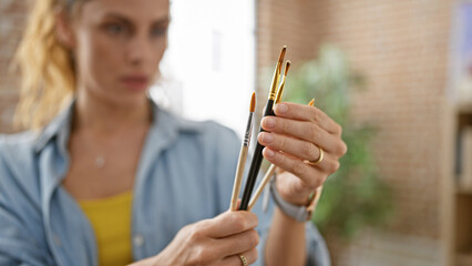 A focused young woman compares paintbrushes in a creative studio, evoking a sense of art, choice, and concentration.