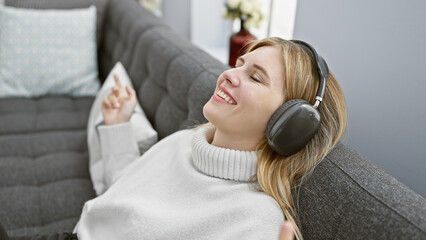 Blonde woman relaxing with closed eyes wearing headphones on a sofa in a cozy living room setting