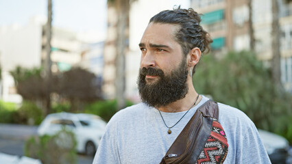 A bearded young man with a trendy hairstyle stands casually on a city street, embodying urban style...