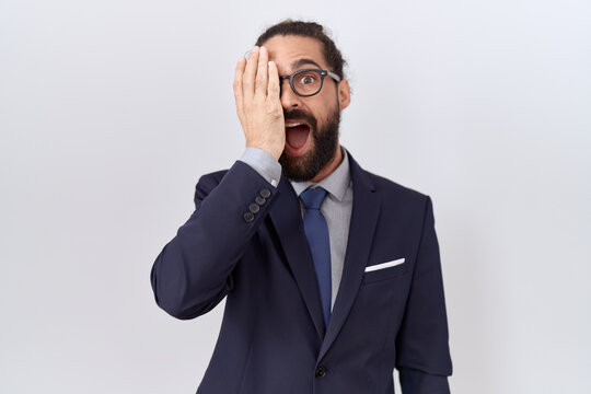 Hispanic man with beard wearing suit and tie covering one eye with hand, confident smile on face and surprise emotion.