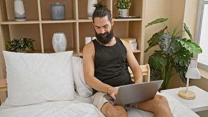 Hispanic man with beard using laptop in well-decorated bedroom.