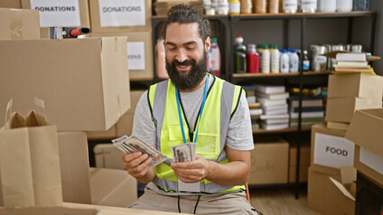 Smiling hispanic man counting money inside a donation center surrounded by cardboard boxes.