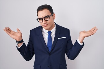 Young hispanic man wearing suit and tie clueless and confused expression with arms and hands raised. doubt concept.