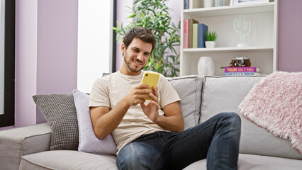 Smiling young hispanic man with a beard using a smartphone while relaxing on a couch in a cozy living room