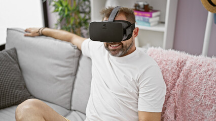 Hispanic mature man enjoys virtual reality headset in modern living room, relaxed on sofa.