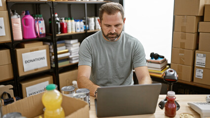Mature man working on laptop in a storage room filled with boxes and donations. - obrazy, fototapety, plakaty