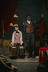 High African man with dirty face standing with little servant boy over dark retro circus backstage background. Artistic performance. Concept of circus, theater, performance, show, retro and vintage