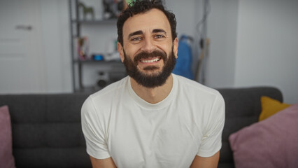 A smiling man with a beard sitting in a modern living room wearing a white shirt