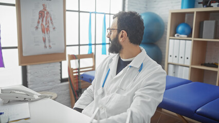 Fototapeta premium A handsome bearded man in a white coat sits thoughtfully in a medical clinic's consulting room.