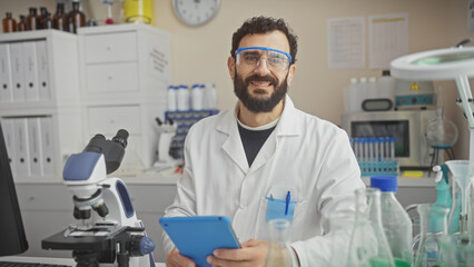 Smiling bearded man in lab coat and safety glasses holding a tablet inside a laboratory filled with...