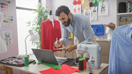A bearded man in a tailor shop measures fabric with sewing tools and a laptop on his worktable.