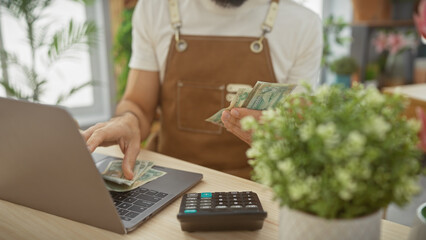 A man in a flower shop counts uae dirhams near a laptop and calculator, indicating business...
