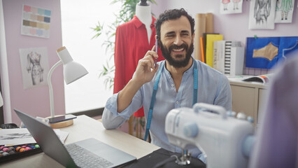 A smiling bearded man in a tailor shop is on the phone, surrounded by sewing equipment, fabric, and...