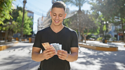 Handsome hispanic man counts mexican pesos outdoors in a sunny city park.