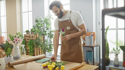 A bearded man in an apron arranges flowers in a sunny, plant-filled flower shop interior.