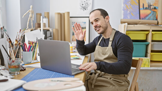 A smiling bearded man wearing an apron gestures during a video call in a bright art studio filled with canvases and supplies.
