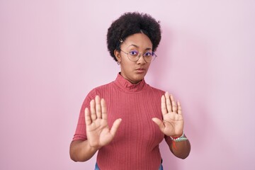 Beautiful african woman with curly hair standing over pink background moving away hands palms...