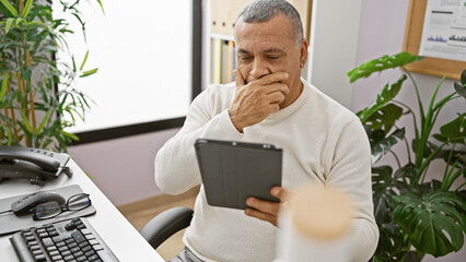 A mature hispanic man in a white sweater uses a tablet in a modern office, displaying concern or...