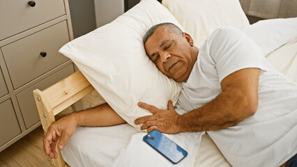 Middle-aged hispanic man sleeping peacefully in a cozy bedroom with a phone beside him