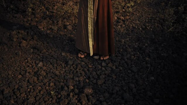 Close-up of a person's feet on a rocky surface, wearing sandals and a flowing brown skirt, with a mysterious and moody atmosphere.