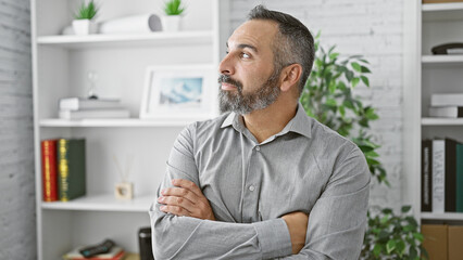 Fototapeta premium Hispanic senior man with beard and grey hair standing arms crossed in a modern office interior.