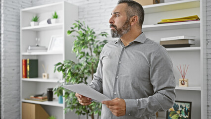 Mature hispanic man with beard and grey hair reading a document in a modern office room.