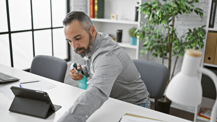 A mature hispanic man with a beard and grey hair is sanitizing his hands in an office interior.