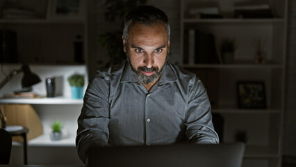 Mature hispanic man with grey beard working late in a dark office indoor setting.