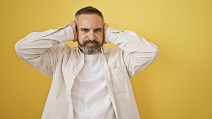 Mature hispanic man with beard frowning and covering his ears against a yellow background.