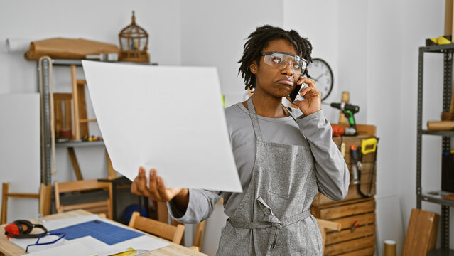 Focused african american woman with dreadlocks wearing safety glasses and apron talks on phone in carpentry workshop.