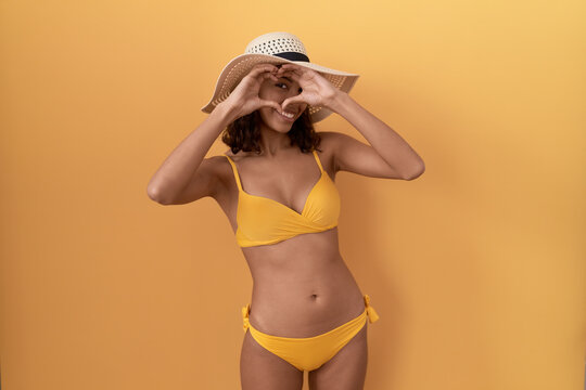 Young hispanic woman wearing bikini and summer hat doing heart shape with hand and fingers smiling looking through sign